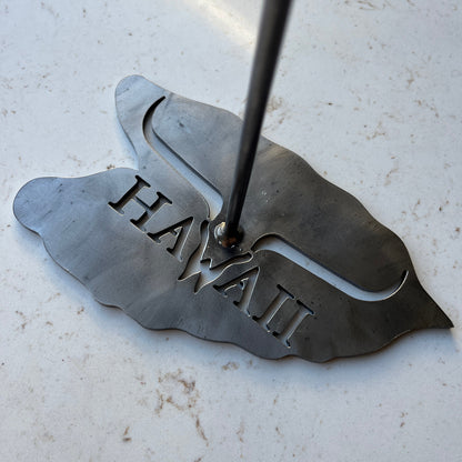 A close-up of a custom steel laser-cut hat stand base, on a light stone or concrete surface.